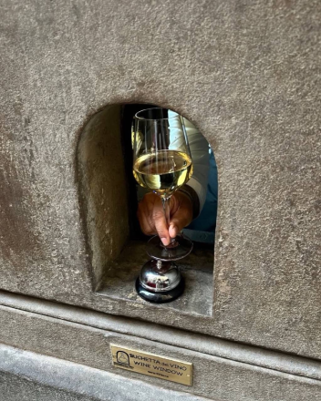 A hand receiving a glass of wine through a tiny stone wine window.